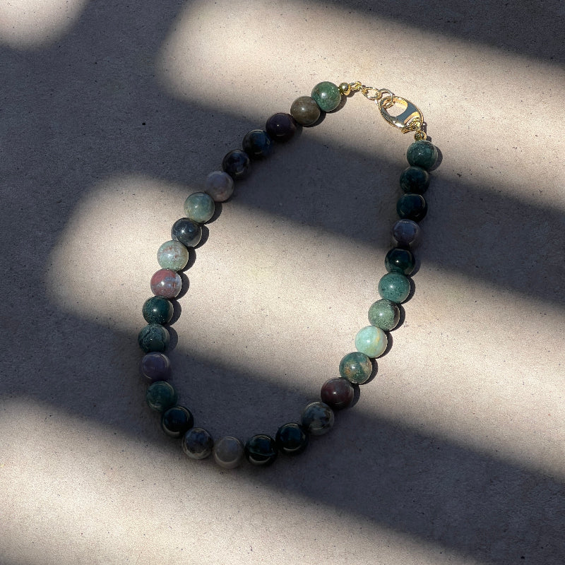 A beaded necklace made of polished round stones in shades of green, blue, gray, and purple, with a small gold clasp, photographed on a light concrete surface with strong diagonal shadows.