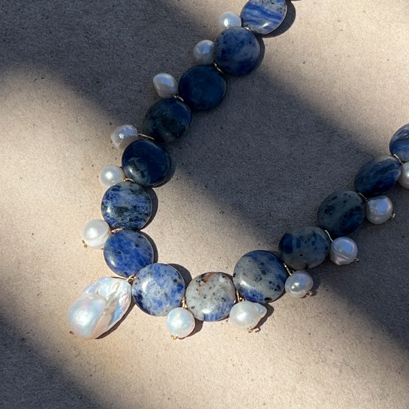 A beaded necklace featuring round and irregular dark blue and white marbled stone beads, accented with small white pearl-like beads, laid out on a textured beige surface with diagonal shadows.