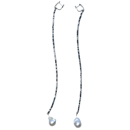A pair of long drop earrings with silver-toned hoops connected by thin beaded chains, each ending in a teardrop-shaped white pearl, displayed on a textured beige fabric background.