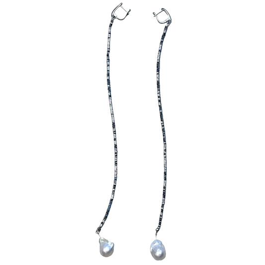 A pair of long drop earrings with silver-toned hoops connected by thin beaded chains, each ending in a teardrop-shaped white pearl, displayed on a textured beige fabric background.