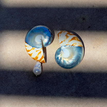 A pair of unique shell-shaped earrings with iridescent blue and white sections, orange marbled accents, and a small white pearl drop, displayed on a concrete surface under direct sunlight.
