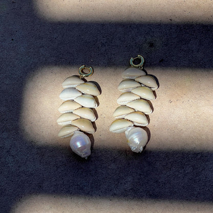 A pair of handmade dangle earrings made of stacked cowrie shells and a large pearl, displayed on a concrete surface under direct sunlight with shadows.
