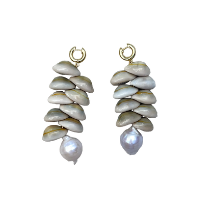 A pair of handmade dangle earrings featuring stacked natural clam shells in beige and gray tones, with a small gold hoop at the top and a single baroque pearl dangling at the bottom, displayed on a textured beige linen background.