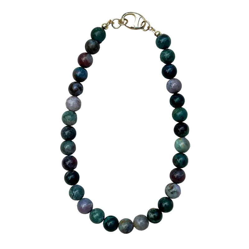 A beaded necklace made of polished round gemstone beads in shades of green, dark purple, and gray, with a gold clasp, laid flat on a textured off-white fabric background.