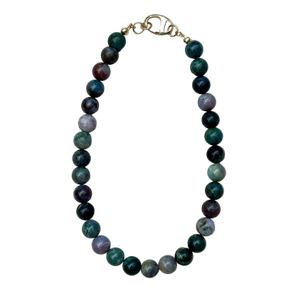 A beaded necklace made of polished round gemstone beads in shades of green, dark purple, and gray, with a gold clasp, laid flat on a textured off-white fabric background.