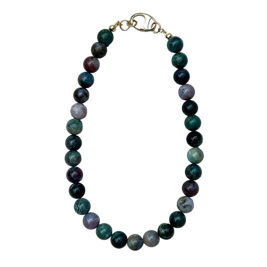 A beaded necklace made of polished round gemstone beads in shades of green, dark purple, and gray, with a gold clasp, laid flat on a textured off-white fabric background.