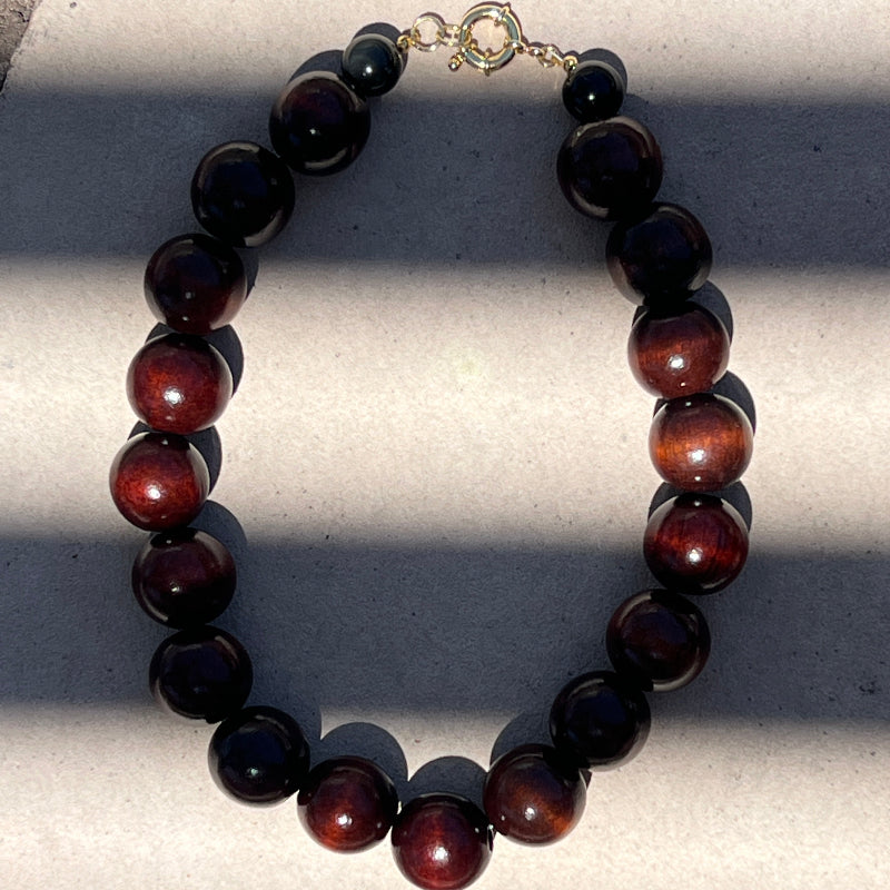 A beaded necklace made of glossy dark brown and black round beads with a small gold clasp, photographed on a light surface with strong diagonal shadows.
