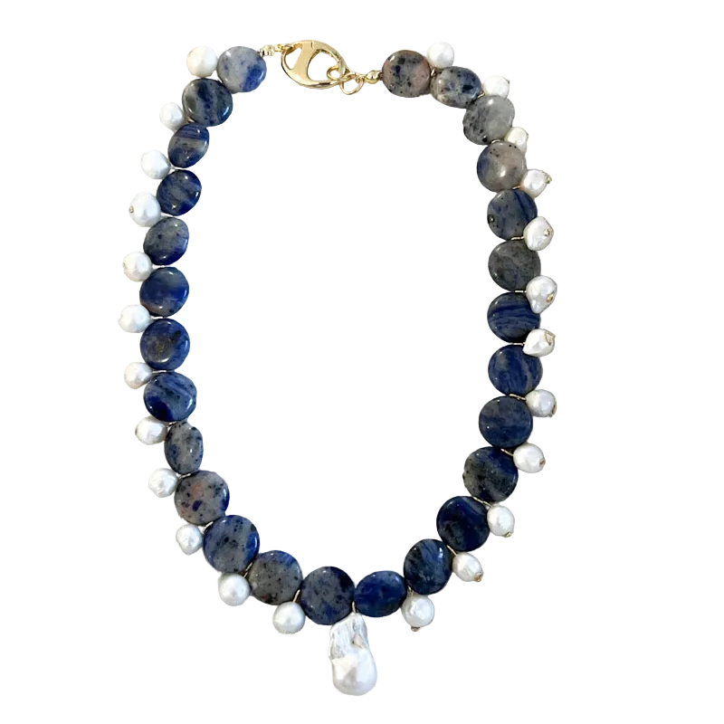 A beaded necklace featuring dark blue sapphire-like beads, small white pearl-like beads, and a gold clasp, displayed on a textured beige fabric background.