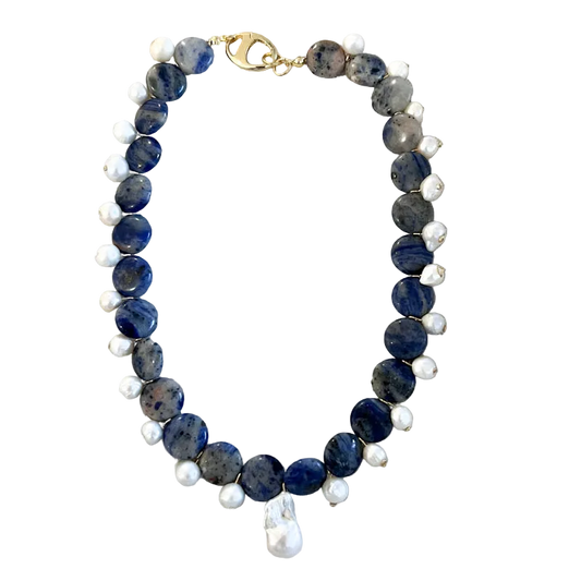 A beaded necklace featuring dark blue sapphire-like beads, small white pearl-like beads, and a gold clasp, displayed on a textured beige fabric background.