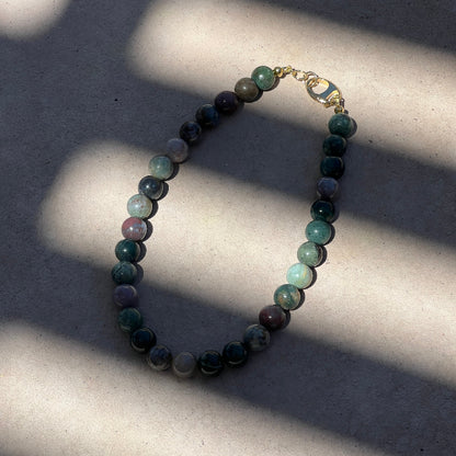 A beaded necklace made of polished round stones in shades of green, blue, gray, and purple, with a small gold clasp, photographed on a light concrete surface with strong diagonal shadows.