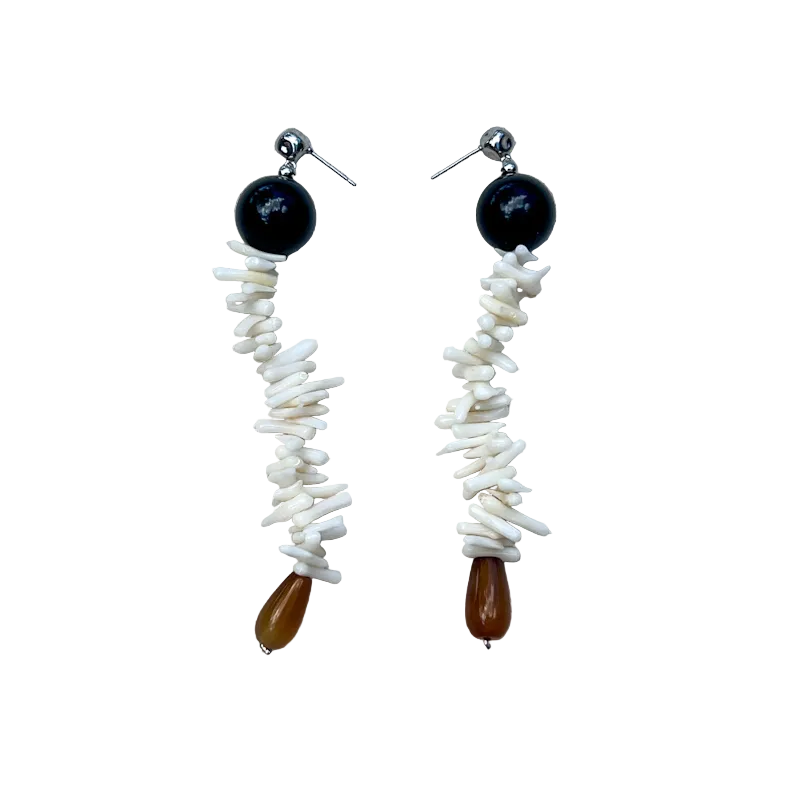 A pair of handmade dangle earrings laid flat on a textured beige linen background. Each earring features a round black bead, a vertical strand of white shell fragments, and a small teardrop-shaped brown bead at the bottom.