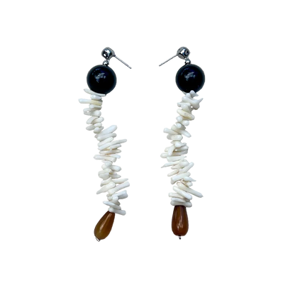 A pair of handmade dangle earrings laid flat on a textured beige linen background. Each earring features a round black bead, a vertical strand of white shell fragments, and a small teardrop-shaped brown bead at the bottom.