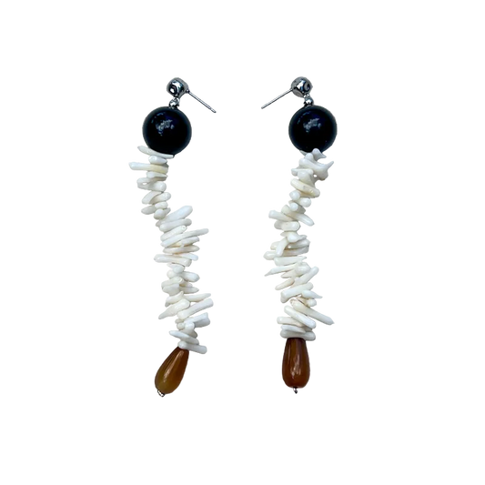 A pair of handmade dangle earrings laid flat on a textured beige linen background. Each earring features a round black bead, a vertical strand of white shell fragments, and a small teardrop-shaped brown bead at the bottom.
