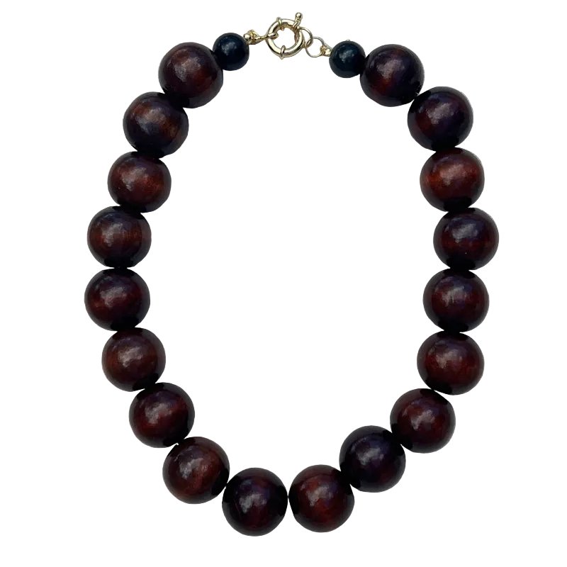 A beaded necklace made of glossy dark reddish-brown wooden beads, accented with two small black beads and a gold-tone clasp, displayed on a light gray textured fabric background.