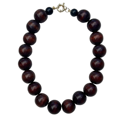 A beaded necklace made of glossy dark reddish-brown wooden beads, accented with two small black beads and a gold-tone clasp, displayed on a light gray textured fabric background.