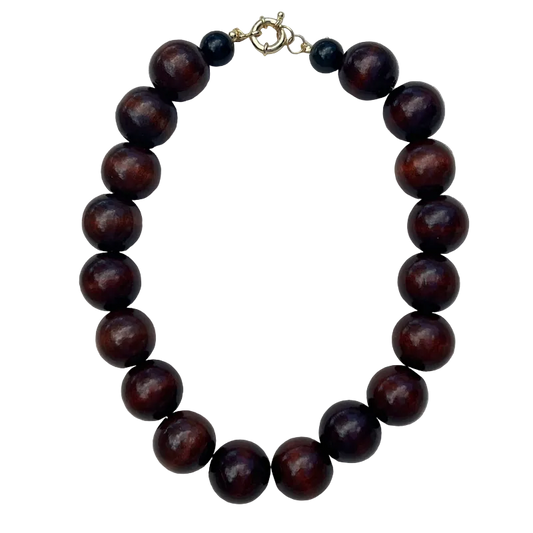A beaded necklace made of glossy dark reddish-brown wooden beads, accented with two small black beads and a gold-tone clasp, displayed on a light gray textured fabric background.